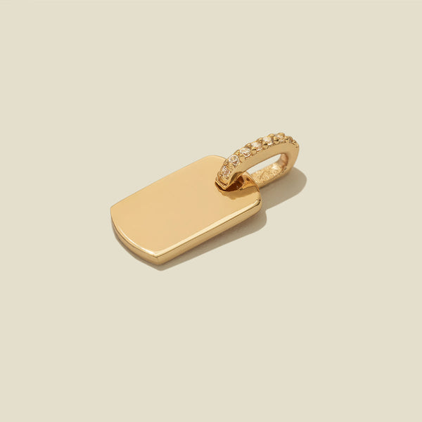 A minimalist gold dog tag pendant with a small row of clear pavé-set stones along the top bail, centered on a solid light beige background.