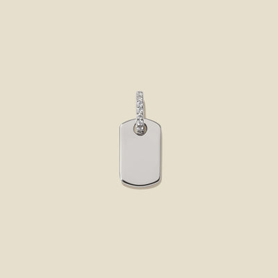 A minimalist silver dog tag pendant with a small row of clear pavé stones along the bail, on a light beige background.