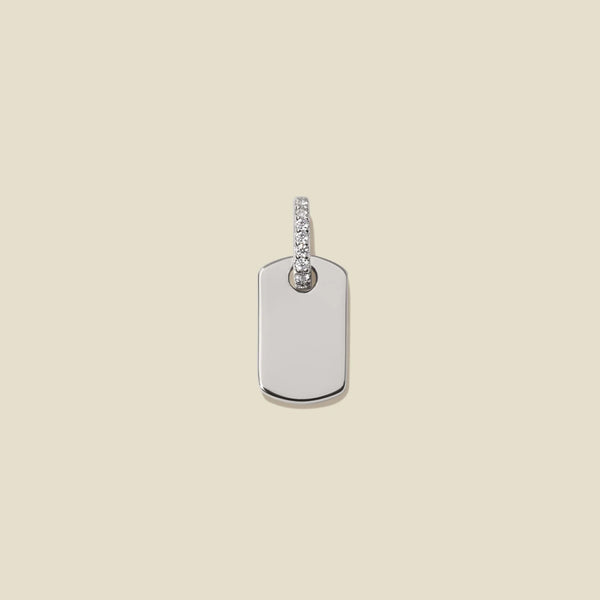 A minimalist silver dog tag pendant with a small row of clear pavé stones along the bail, on a light beige background.