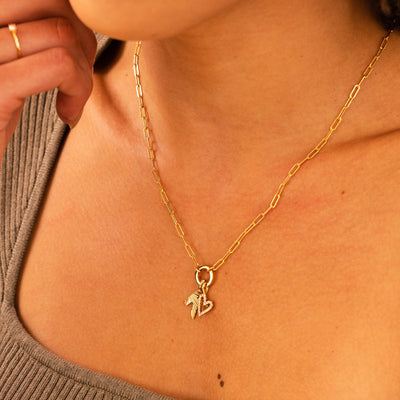 A person wearing a gold paperclip link chain necklace with a round ring in the center, holding an initial 'K' charm and a heart charm embellished with clear accent stones.