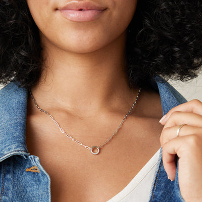 A close-up of a person's chest wearing a silver paperclip link chain necklace with a round silver ring in the center, embellished with clear accent stones.