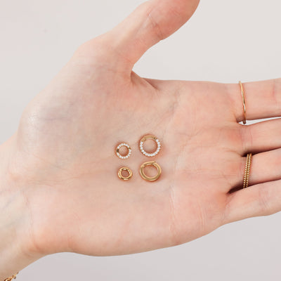 A person's open hand holding four round gold round ring attachments in two different sizes. One set is a polished gold and the other is covered in small clear accent stones.