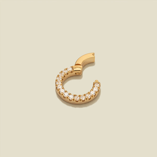A small gold ring necklace attachment embellished with a row of clear pavé-set stones, shown with the latch open on a solid beige background.