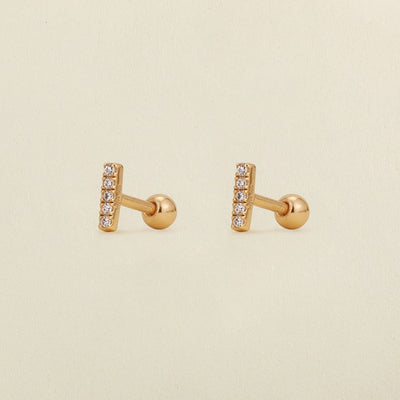 A pair of gold stud earrings with a vertical bar set with clear cubic zirconia stones, shown on a light beige textured background.
