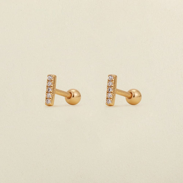 A pair of gold stud earrings with a vertical bar set with clear cubic zirconia stones, shown on a light beige textured background.