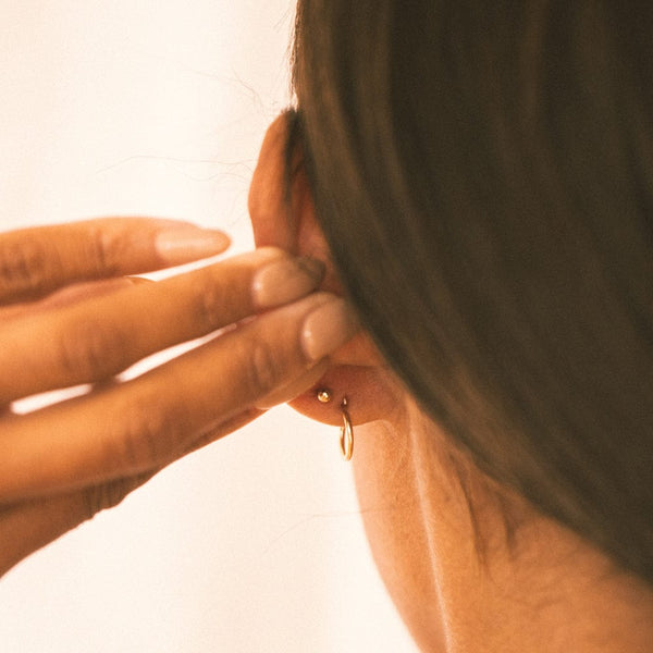 The back of a person's ear wearing a thin gold hoop earring and a gold round ball backing for a stud.
