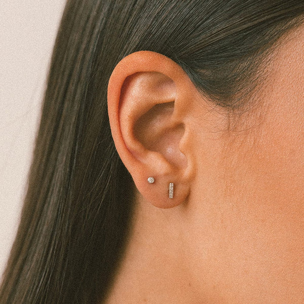 A person's ear wearing a silver vertical bar stud with clear gemstones and a small silver stud with a bezel-set clear gemstone.