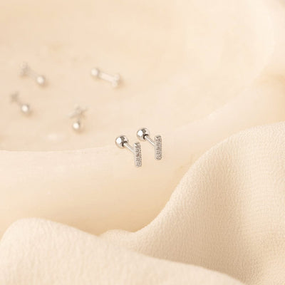 A pair of silver stud earrings with a vertical bar set with clear cubic zirconia stones, shown on a textured cream colored wavy surface with other silver studs blurred in the background.