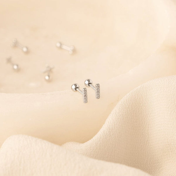 A pair of silver stud earrings with a vertical bar set with clear cubic zirconia stones, shown on a textured cream colored wavy surface with other silver studs blurred in the background.