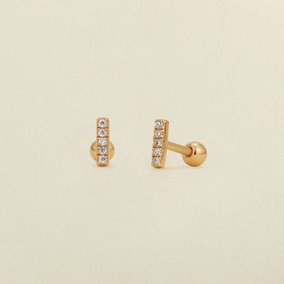 A pair of gold stud earrings with a vertical bar set with clear cubic zirconia stones, shown on a light beige textured background.