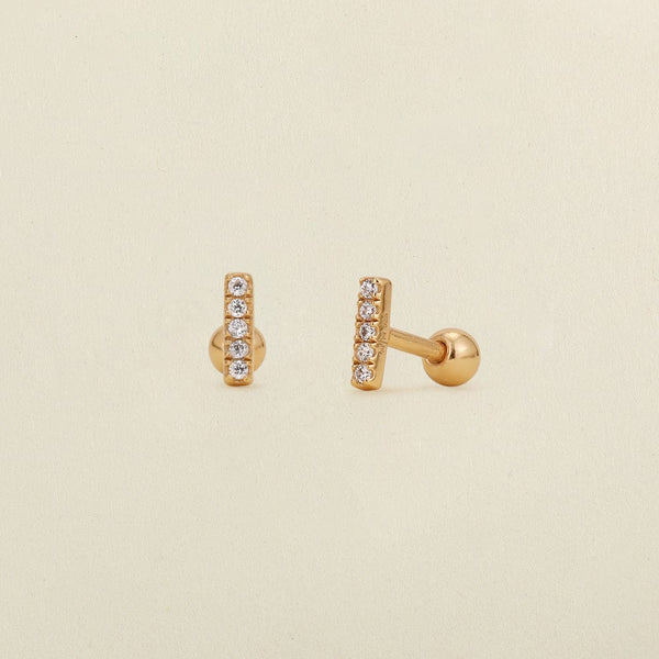 A pair of gold stud earrings with a vertical bar set with clear cubic zirconia stones, shown on a light beige textured background.