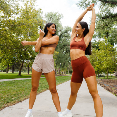 Two women in athletic apparel on a sidewalk in the park, stretching their arms.