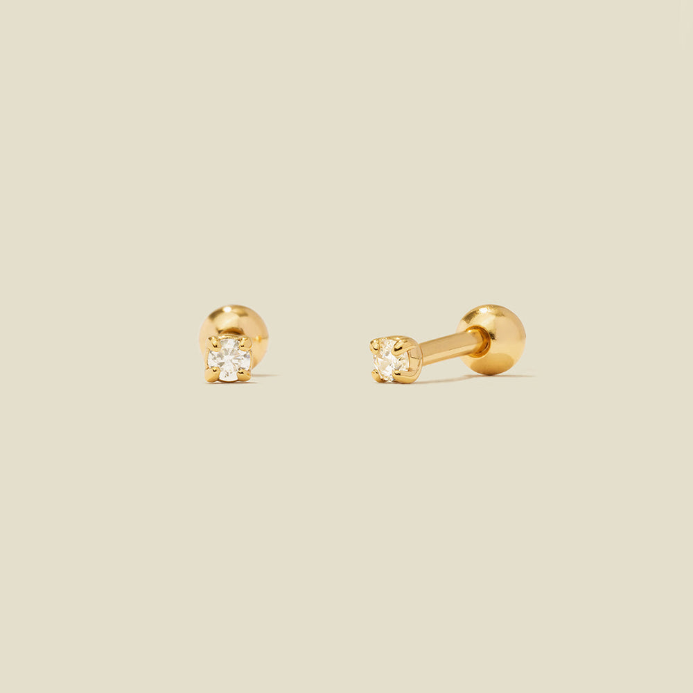 A pair of gold stud earrings with round prong-set clear gemstones, shown on a solid light beige background.