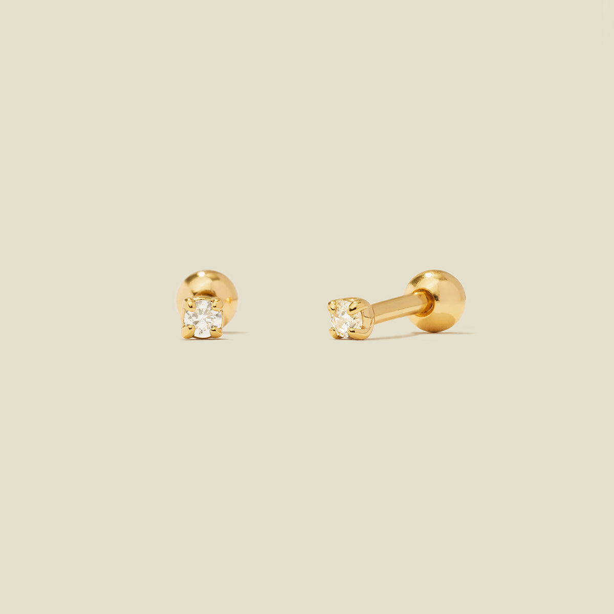 A pair of gold stud earrings with round prong-set clear gemstones, shown on a solid light beige background.