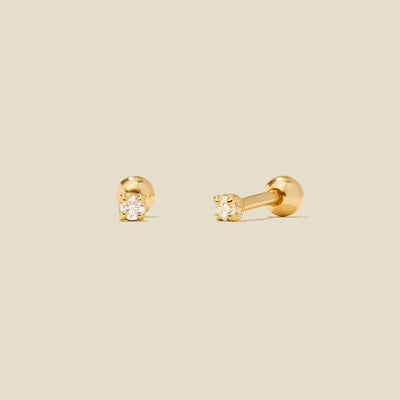 A pair of gold stud earrings with round prong-set clear gemstones, shown on a solid light beige background.
