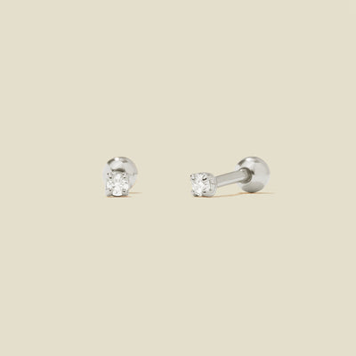A pair of silver stud earrings with round prong-set clear gemstones, shown on a solid light beige background.
