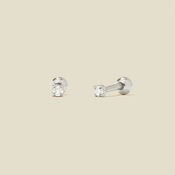 A pair of silver stud earrings with round prong-set clear gemstones, shown on a solid light beige background.