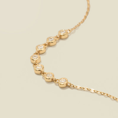 A gold delicate chain bracelet with seven round bezel-set clear gemstones in a row, laying on a solid beige surface.