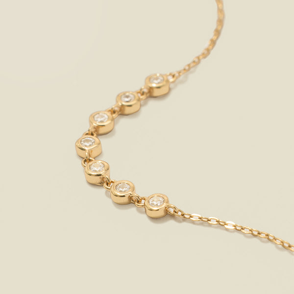 A gold delicate chain bracelet with seven round bezel-set clear gemstones in a row, laying on a solid beige surface.