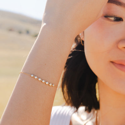 A close up of a woman holding her hand up to her face, showcasing a delicate gold chain bracelet with a short section of small, round bezel-set clear gemstones side by side.