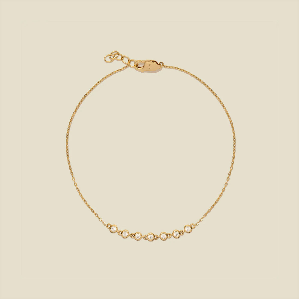 A delicate gold chain bracelet with a short section of small, round bezel-set clear gemstones side by side at the bottom, displayed on a solid light beige background.