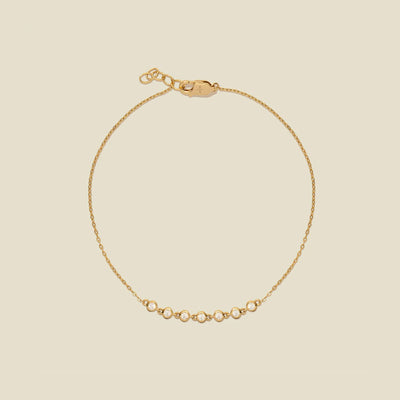 A delicate gold chain bracelet with a short section of small, round bezel-set clear gemstones side by side at the bottom, displayed on a solid light beige background.