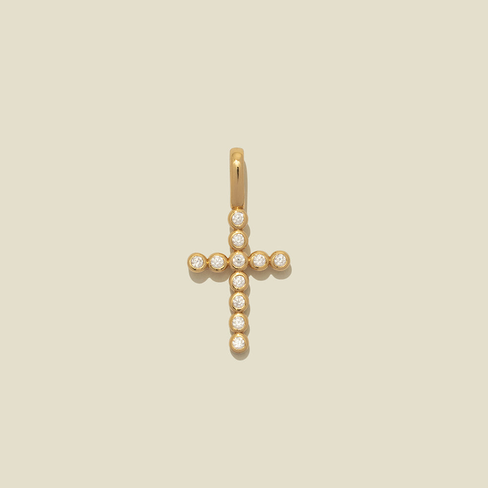 A gold cross pendant adorned with small clear gemstones, centered on a solid light beige background.