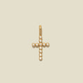 A gold cross pendant adorned with small clear gemstones, centered on a solid light beige background.