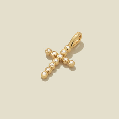 A gold cross charm pendant comprised of small round bezel-set clear gemstones, laying at an angle on a solid beige surface.