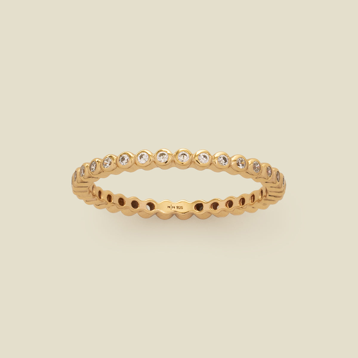 A gold eternity ring featuring a continuous row of bezel-set clear round gemstones on a light beige background.