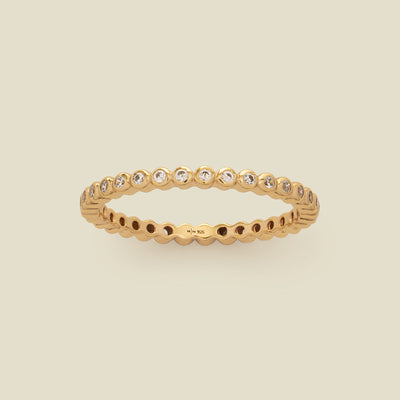 A gold eternity ring featuring a continuous row of bezel-set clear round gemstones on a light beige background.
