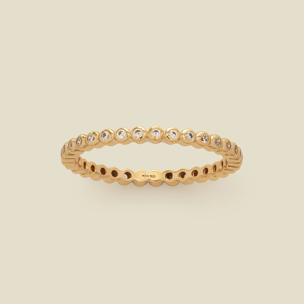 A gold eternity ring featuring a continuous row of bezel-set clear round gemstones on a light beige background.