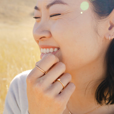 A close-up of a woman with her hand to her face, showcasing a gold thin band ring with a single pearl and a gold eternity style ring made up of small round bezel-set clear gemstones, standing in front of a beige field blurred in the background.