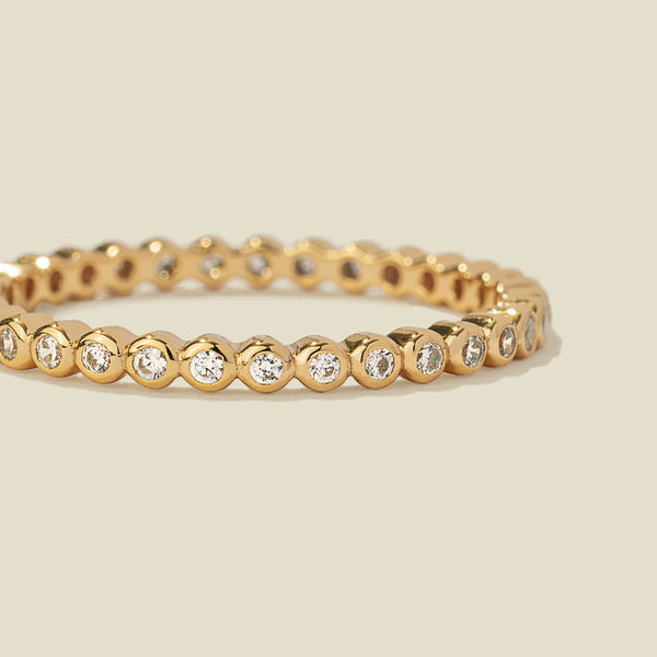 A gold eternity ring featuring a continuous row of bezel-set clear round gemstones on a light beige background.