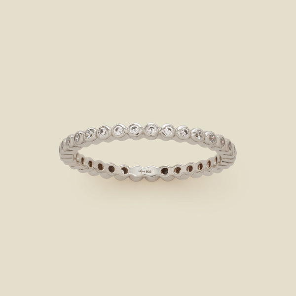 A silver eternity ring featuring a continuous row of bezel-set clear round gemstones on a light beige background.