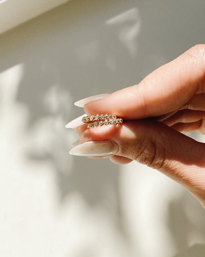 A person's hand holding two gold earrings featuring round bezel-set clear gemstones, with a shadowy, white background.