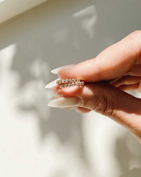 A person's hand holding two gold earrings featuring round bezel-set clear gemstones, with a shadowy, white background.