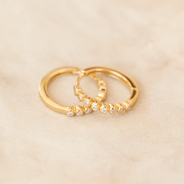 A pair of gold hoop earrings featuring round bezel-set clear gemstones, laying on top of one another on a marbled, beige background.