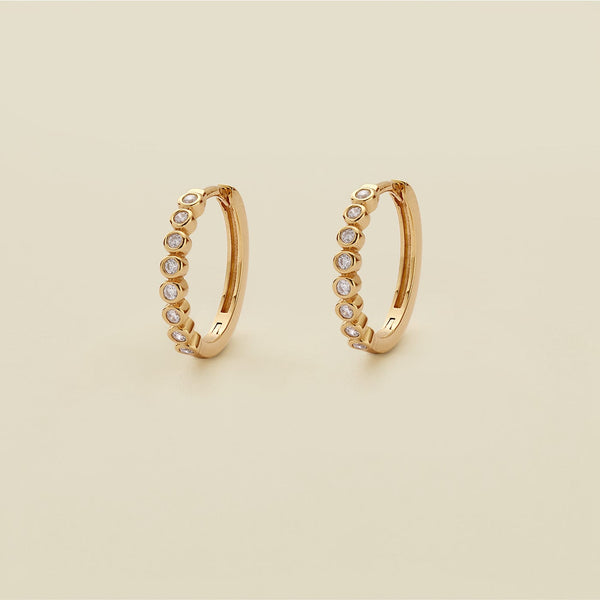 A pair of gold hoop earrings featuring round bezel-set clear gemstones, standing upright against a solid beige background.