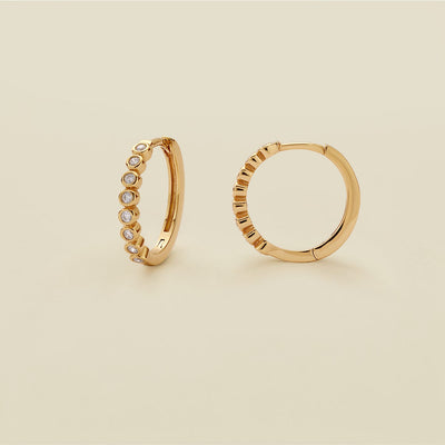A pair of gold hoop earrings featuring round bezel-set clear gemstones, standing upright against a solid beige background.
