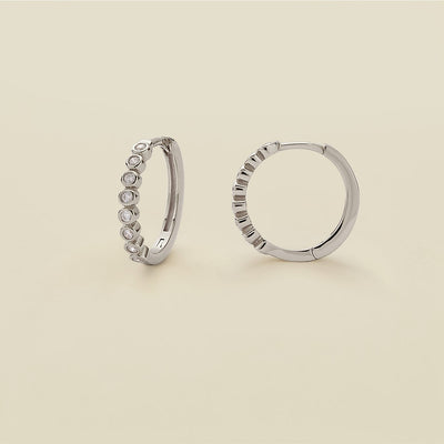 A pair of silver hoop earrings featuring round bezel-set clear gemstones, standing upright against a solid beige background.
