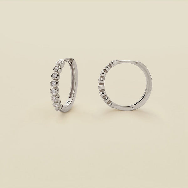A pair of silver hoop earrings featuring round bezel-set clear gemstones, standing upright against a solid beige background.