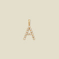 A gold letter 'A' pendant charm embellished with small clear round gemstones, centered on a solid light beige background.