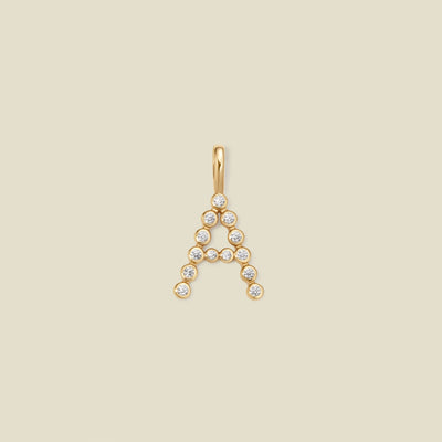 A gold letter 'A' pendant charm embellished with small clear round gemstones, centered on a solid light beige background.