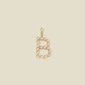 A gold letter 'B' pendant charm embellished with clear round gemstones, centered on a solid light beige background.