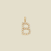 A gold letter 'B' pendant charm embellished with clear round gemstones, centered on a solid light beige background.