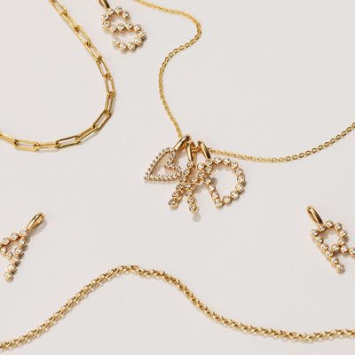 Three gold chains in various styles laying on a white background with multiple gold initial charms and a gold heart charm, all with clear gemstone accents.