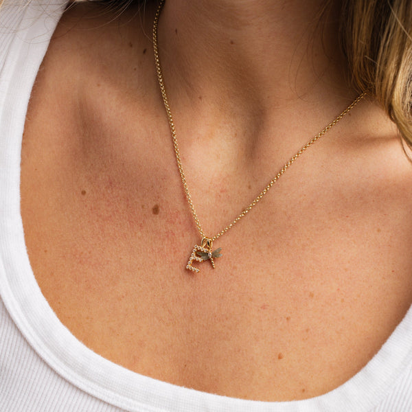 A person wearing a gold chain necklace with an initial 'E' charm and a gold dragonfly charm with natural stones on the wings, paired with a white ribbed top.