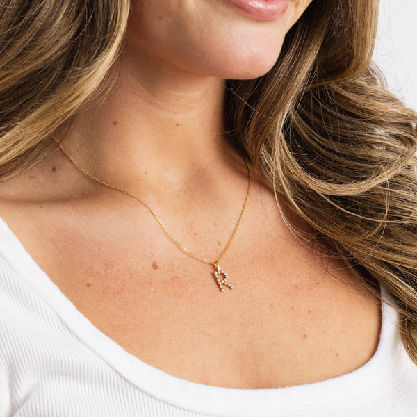 A close-up of a woman wearing a gold pendant necklace with a gold initial 'R' charm, paired with a white ribbed top and set against a solid white background.