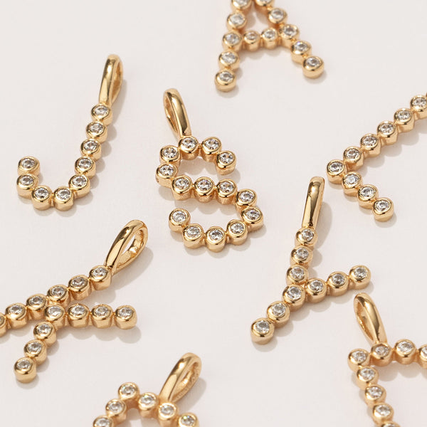 Multiple gold initial charms comprised of clear round bezel-set gemstones, scattered on a solid white surface with the letters 'J', 'S', and 'Y' fully visible.